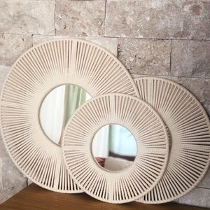 May include: Three round mirrors with a woven rope frame. The mirrors are nested inside each other, with the largest mirror in the back and the smallest mirror in the front. The mirrors are all cream colored.