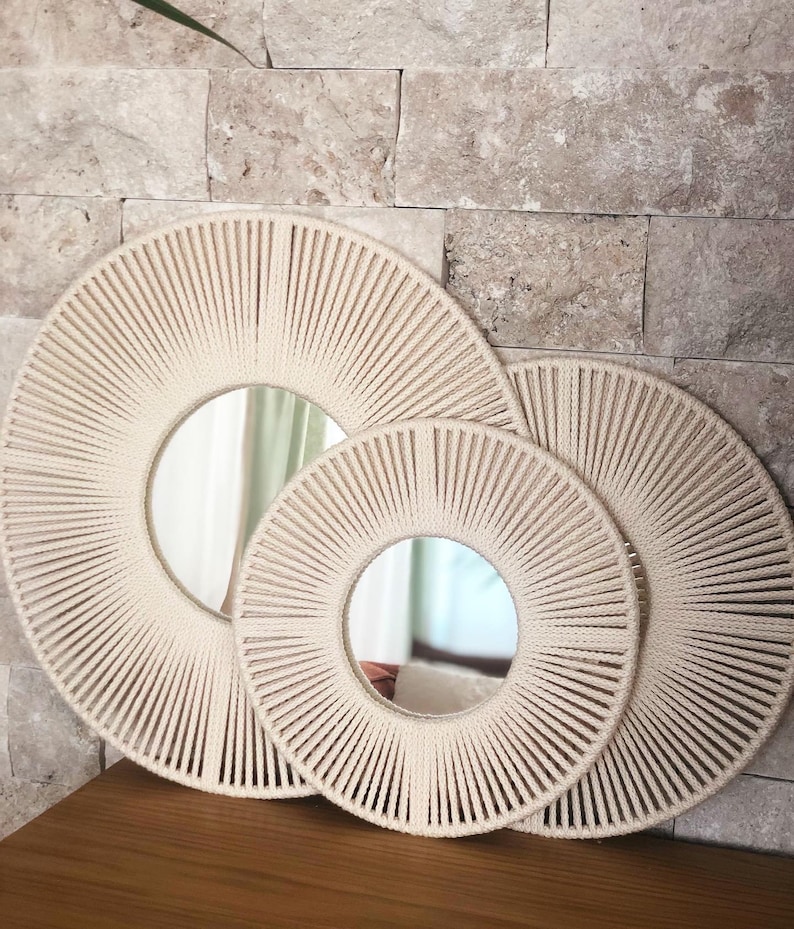 May include: Three round mirrors with a woven rope frame. The mirrors are nested inside each other, with the largest mirror in the back and the smallest mirror in the front. The mirrors are all cream colored.