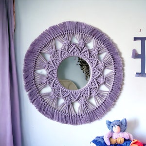 May include: A circular macrame mirror in lavender, featuring a central mirror and a detailed knotted design with a fringed border. The mirror is mounted on a white wall. The overall diameter is approximately 50 cm.