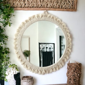 May include: A round macrame mirror with a white fringe border. The mirror is hanging on a white wall above a wooden dresser.