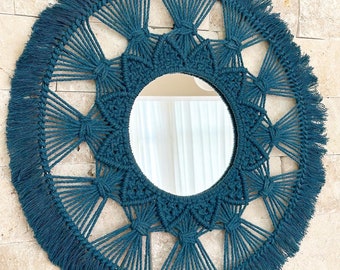 May include: A teal blue macrame mirror with a circular shape and a fringe border. The mirror has a geometric design with a mandala-like pattern.
