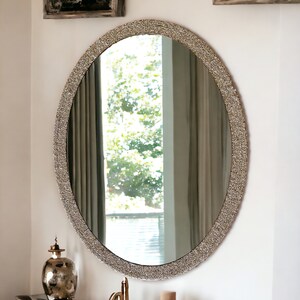 Puede incluir: Un gran espejo ovalado con un marco decorativo, brillante, plateado. El espejo est&aacute; colgado en una pared blanca sobre un tocador blanco con un grifo dorado.