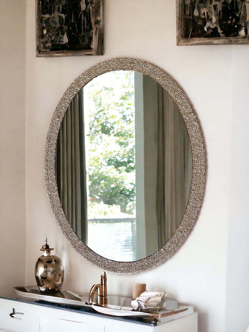 Puede incluir: Un gran espejo ovalado con un marco decorativo, brillante, plateado. El espejo est&aacute; colgado en una pared blanca sobre un tocador blanco con un grifo dorado.