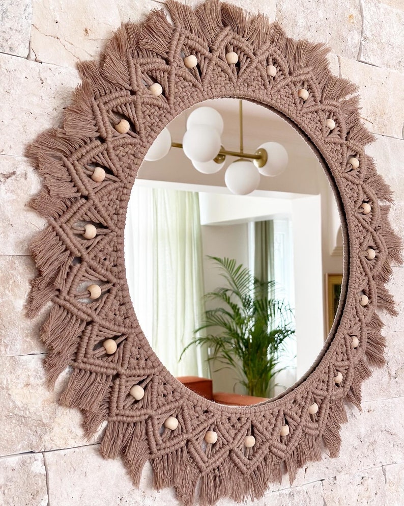 May include: A round mirror with a brown macrame frame and wooden beads. The frame has a fringe around the edge.
