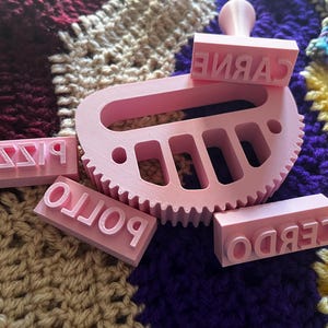 May include: Pink 3D-printed stamps with the words "Pizza", "Pollo", "Cerdo", and "Carne" in reverse. A pink gear-shaped tool is also present. The items are on a colorful crocheted blanket. Ideal for crafting or cooking.