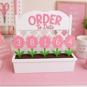 May include: A white wooden order tracker with pink lettering that reads "ORDER To Date." Five pink flower cutouts display the numbers 3, 8, 1, 6, and 4. The tracker sits in a white planter box with brown soil.