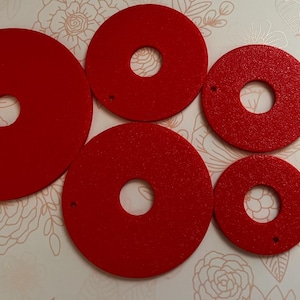 May include: A collection of red, circular felt pieces with a hole in the center. The pieces vary in size, ranging from large to small. The felt has a textured surface. The objects are arranged on a floral patterned background.