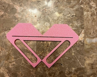 May include: A pink heart-shaped craft with two cut-out ovals in the center. The craft is made of foam or cardboard and is sitting on a marble surface.