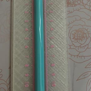 May include: A light blue pen with a silver tip rests on a clear plastic ruler. The ruler has pink markings and numbers from 0 to 13 inches. The background features a floral pattern.