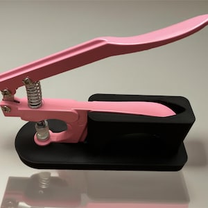 May include: A pink hand press tool with a black base. The tool has a long handle and a spring mechanism. The tool is designed for applying pressure. The tool is on a white surface.
