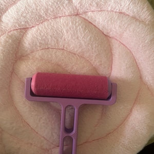 May include: A magenta paint roller with a purple handle rests on a light pink, plush, circular surface. The roller is made of a dense, cylindrical foam. The handle has a T-shape with two oval cutouts. The plush surface has a spiral design.