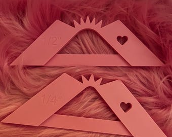 May include: Two pink quilting guides with a heart-shaped cutout and a sunburst design. One guide is labeled "1/2" and the other "1/4". The guides are made of a solid material and are set against a pink, fluffy background.