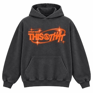 Peut inclure: Un sweat à capuche gris anthracite avec une poche kangourou sur le devant et un motif imprimé orange vif. Le design comprend le texte STRAY KIDS et THIS & THAT entouré d'une forme ovale et d'accents en forme d'étoiles à quatre branches.