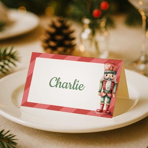 Op de afbeelding: Een feestelijke naamkaart met de naam "Charlie" in groen schrift, omlijst door een rood-witte gestreepte rand. Een aquarel illustratie van een notenkraker soldaat versiert de rechterkant. De kaart ligt op een wit bord, onderdeel van een feestelijke tafelsetting.