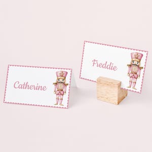 Op de afbeelding: Twee witte tafelkaarten met roze geschulpte randen en de namen "Catherine" en "Freddie" in roze script. Elke kaart heeft een illustratie van een roze notenkrakersoldaat. Een houten blok houdt een kaart vast.