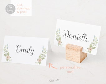 May include: Two white place cards with floral designs and the names "Emily" and "Danielle" written in black cursive. The cards are on a white surface with a wooden stand holding one of the cards.