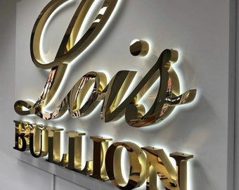 May include: Gold-colored metal sign with the text "Lois Bullion" in a cursive font. The letters are raised and have a halo of light around them.