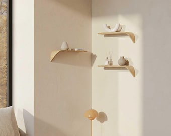 May include: Three wooden floating shelves with a curved design are mounted on a beige wall. Each shelf holds decorative items like vases and books. A small lamp sits on a side table in the foreground, adding to the room's minimalist aesthetic.