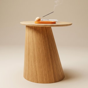 Op de afbeelding: Een bijzettafel van lichtgekleurd hout met een conische voet en een rond tafelblad. Op de tafel staat een oranje rechthoekig dienblad met een houten bolvormige wierookhouder en een brandend wierookstokje dat rook afgeeft.
