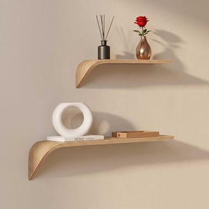 Peut inclure: Deux étagères flottantes en bois incurvées sont fixées au mur beige. L'étagère supérieure contient un diffuseur noir, un vase cuivré avec une rose rouge. L'étagère inférieure affiche un anneau en céramique blanc et un livre avec le mot "HOME".