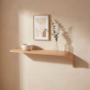 Puede incluir: Una estantería flotante de madera clara con un extremo curvado está montada en una pared beige. Un cuenco blanco, una impresión botánica enmarcada y un jarrón de cristal transparente con vegetación se exhiben en la estantería.