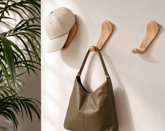 May include: A beige baseball cap rests on a wooden wall-mounted shelf. A brown leather handbag hangs from a wooden hook below. Two additional wooden hooks are also mounted on the wall. The wall is painted white.