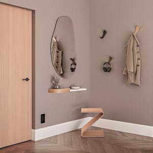 May include: A modern entryway with a large, irregularly shaped mirror, a floating shelf, and a wooden Z-shaped side table. A tan coat hangs on a wooden hook. The walls are painted a neutral color, and the floor has a herringbone pattern.
