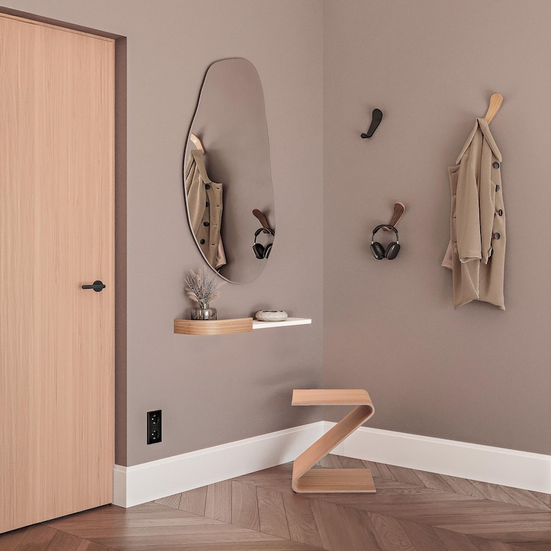 May include: A modern entryway with a large, irregularly shaped mirror, a floating shelf, and a wooden Z-shaped side table. A tan coat hangs on a wooden hook. The walls are painted a neutral color, and the floor has a herringbone pattern.