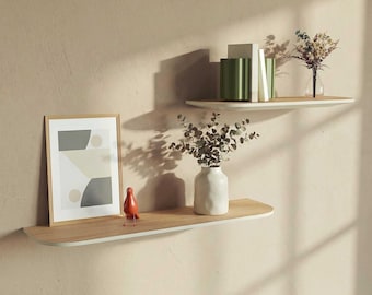 May include: Two floating shelves with a light wood finish and white trim are mounted on a beige wall. The shelves display a framed abstract print, a small orange bird figurine, a white vase with greenery, and books and vases.