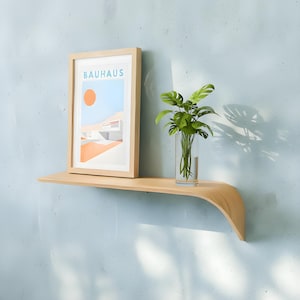 Könnte beinhalten: Ein schwebendes Regal aus hellem Holz mit geschwungener Kante an einer hellblauen Wand. Auf dem Regal befinden sich ein gerahmter "BAUHAUS" Druck und eine Glasvase mit grünen Pflanzen.