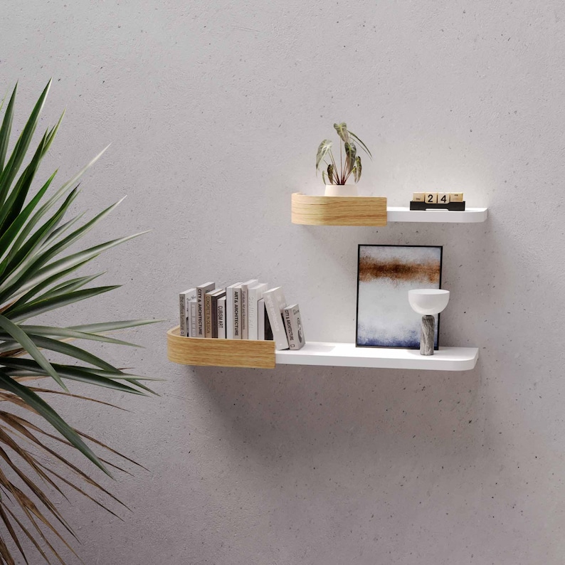 May include: Two-tiered white floating shelves with curved wooden accents. The shelves hold books, a small potted plant, a framed artwork, and a decorative vase. A wooden block calendar displays the date.