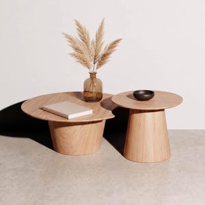 Peut inclure: Deux tables basses en bois clair avec des plateaux circulaires et des bases coniques. Un vase en verre ambré contenant des herbes de la pampa séchées, un livre beige fermé et un petit bol marron foncé sont posés sur les surfaces des tables.