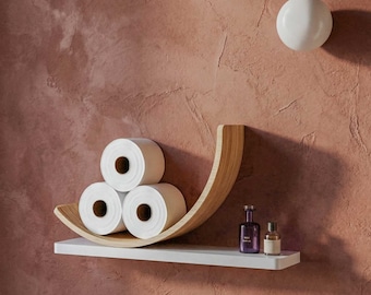 May include: A modern bathroom shelf with three rolls of white toilet paper. The shelf has a curved, light wood design and sits on a white base. Two small bottles are on the shelf. A white, round light fixture is on the wall.