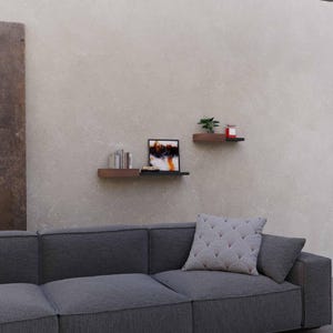 May include: Two floating shelves with a dark wood finish are mounted on a neutral wall. The shelves hold decorative items, including books, a framed abstract painting, and a small potted plant. A gray sofa with a patterned pillow is in the foreground.