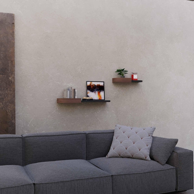 May include: Two floating shelves with a dark wood finish are mounted on a neutral wall. The shelves hold decorative items, including books, a framed abstract painting, and a small potted plant. A gray sofa with a patterned pillow is in the foreground.