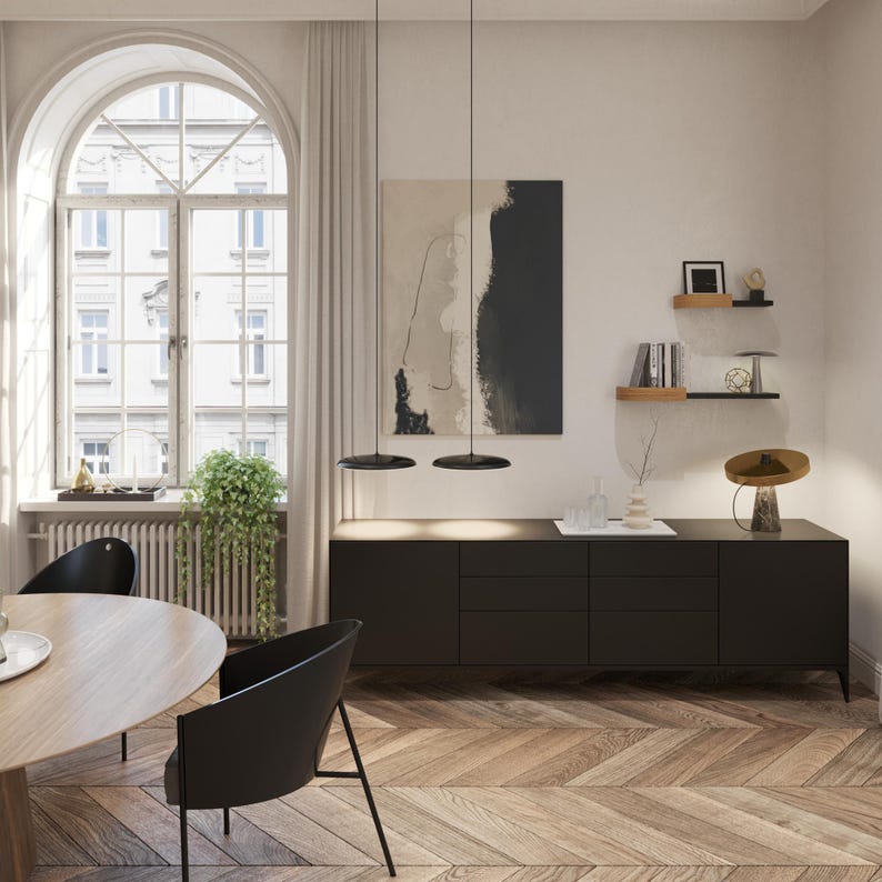 May include: An interior design scene featuring a black sideboard with drawers, a round dining table, and black chairs. A large arched window with a view of a building is visible. The room has a modern aesthetic with a neutral color palette.