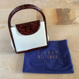Könnte beinhalten: Eine weiße Handtasche mit einer braunen Schildpattklappe und einem oberen Henkel. Die Tasche liegt auf einem blauen Staubbeutel mit dem aufgedruckten Text "Stuart Weitzman".