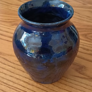 May include: A bulbous ceramic vase with a glossy, deep cobalt blue glaze. The vessel features a wide, rounded body that tapers toward a narrow base and a flared, circular rim. The surface reflects light, showing variations in the blue color intensity.