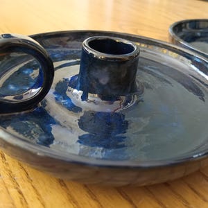 May include: A round, dark blue ceramic candle holder with a glossy finish. The piece features a central cylindrical socket for a candle and a circular side handle for carrying. The surface has subtle variations in blue and black tones.