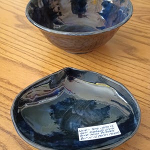 May include: A dark blue glazed ceramic bowl and a matching shell-shaped dish. The dish features a white paper label with the handwritten text: NOTE - THIS ITEM IS NOT MARKED SOUTH BLUE HILLS POTTERY BUT IT IS FROM THEM.
