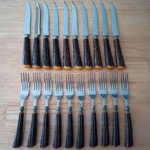 May include: A set of ten silver-toned steak knives and ten silver-toned forks arranged in two rows. Each utensil features a dark brown, textured handle with a distinct vertical grooved pattern and a rounded, light-colored base at the end.