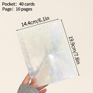May include: A clear, iridescent card holder with a heart pattern, held by a person. The holder is 14.4cm/6.1in wide and 19.9cm/7.8in tall, with 40 card pockets across 10 pages. Text on the image reads "Pocket: 40 cards Page: 10 pages."