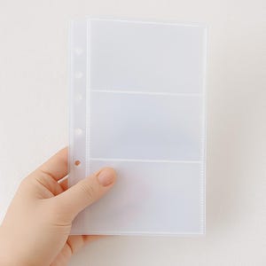 May include: A clear, three-pocket photo album page with a three-hole punch for binder storage. The page is held by a hand against a white background. The pockets are divided by horizontal lines.