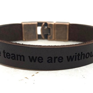 May include: A black leather bracelet with a silver clasp. The bracelet has the text "We be the team we are without you" engraved on it.
