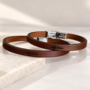 May include: Two brown leather bracelets featuring a small black cross symbol engraved on the surface. Each bracelet is equipped with a silver-toned stainless steel clasp mechanism that secures the band in a circular shape.