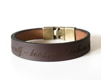 Puede incluir: Pulsera de cuero marrón con cierre plateado. La pulsera tiene las palabras "cree en ti mismo - porque creo en ti" grabadas.
