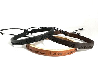 May include: Three leather bracelets with adjustable string closures. The bracelets are brown, black, and tan. Each bracelet has the words "Love Intentionally" and "Purposefully" engraved on them.