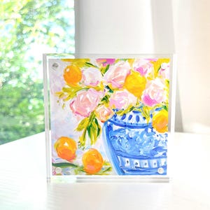 May include: A square acrylic block featuring a colorful floral painting. The artwork depicts a blue and white vase filled with pink and yellow flowers and oranges. The painting has a bright, cheerful aesthetic.