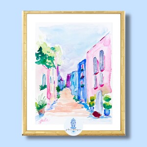 以下が含まれることがあります： カラフルな家と青い空が描かれた水彩画の街並み。家はピンク、青、緑の色合いで描かれています。通りは木々や茂みで縁取られています。絵は金色の額縁に入っています。