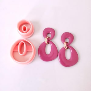 May include: Two pairs of pink resin earrings with a gold accent. The earrings are shaped like a figure eight with a smaller circle in the center. The earrings are shown next to two pink plastic cookie cutters shaped like a figure eight.
